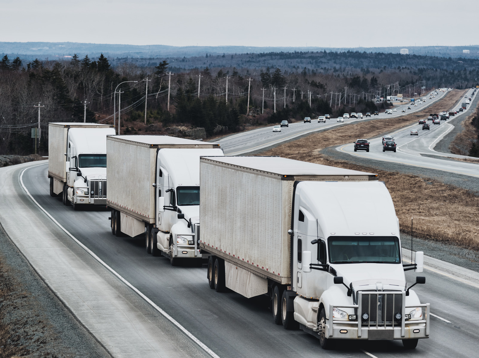 Three white semi-trucks, coordinated with precision akin to a freight brokerage team, drive in a line on the highway. Trees form a serene backdrop, while cars move steadily in the opposite lane.