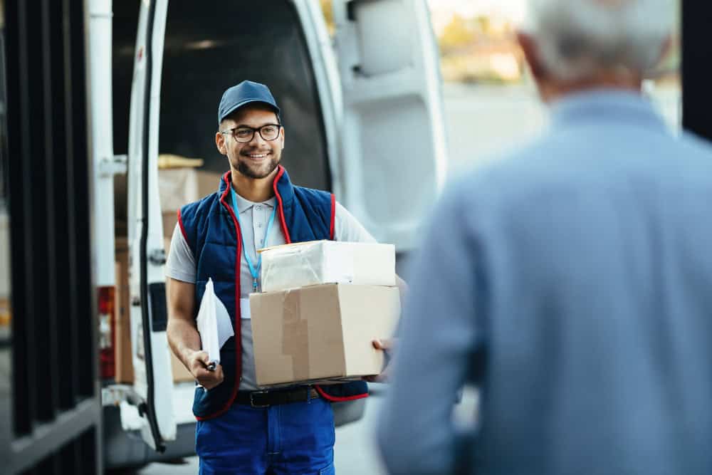 A delivery person grins as they handle packages and paperwork near a van, embodying the seamless coordination of freight brokerage.