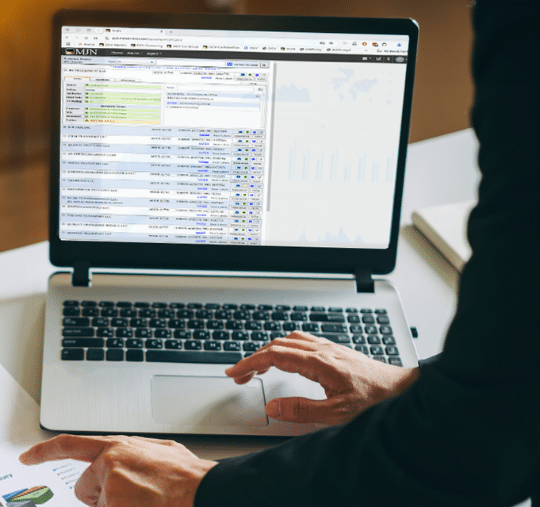A person uses a laptop showing financial management software filled with data and graphs, pointing at a paper with additional charts on the desk, while considering critical factors before choosing a Freight Factoring Company.
