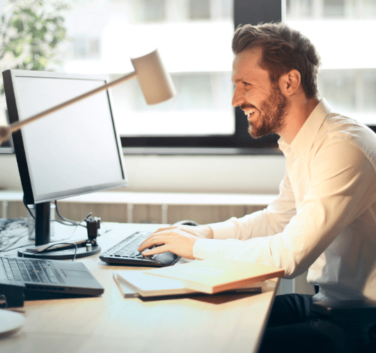A man in a white shirt sits at a desk with two laptops and a book, smiling as he explores critical factors of choosing a freight factoring company, all within the bright setting of his office.