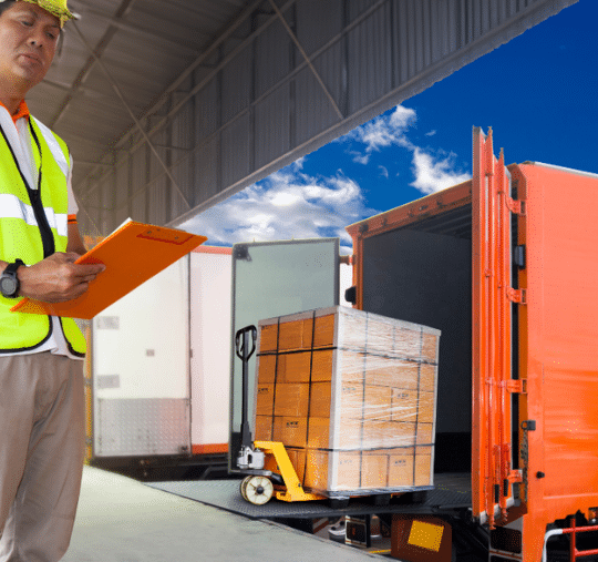 A worker in safety gear holds a clipboard near an orange truck being loaded with boxes on a pallet jack, carefully choosing critical factors to ensure the freight factoring company's protocols are followed in the bustling warehouse.