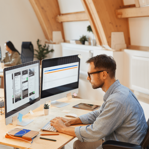 Man working at a desk with two monitors in a modern office; one screen shows a design interface while the other displays a dashboard analyzing trucking factoring costs. Another person works in the background.